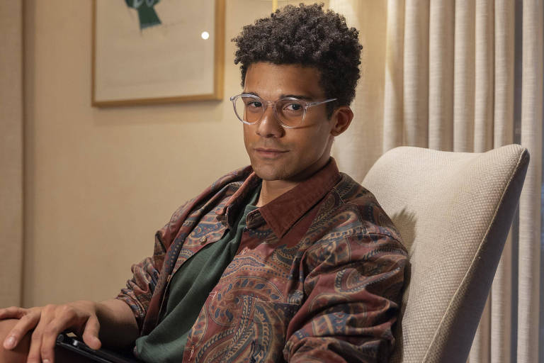 app pagando no cadastro via pix - Um jovem sentado em uma cadeira confortável, usando óculos e uma camisa de manga curta verde sob uma jaqueta estampada. Ele tem cabelo cacheado e está olhando para a câmera com uma expressão neutra. Ao fundo, há cortinas claras e uma parede com uma obra de arte emoldurada.