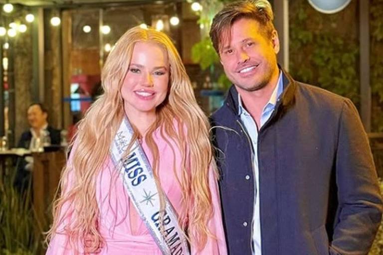 iwcpg.cc - Miss Grand Brasil 2023 veste vestido longo rosa e faixa, segurando clutch branca, ao lado de homem com casaco azul escuro em ambiente iluminado e decorado.