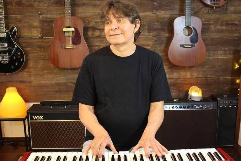 isadora vale nude - Homem de camiseta preta toca teclado em estúdio com parede de madeira ao fundo. Três guitarras penduradas na parede, amplificadores e uma luminária amarela ao lado.