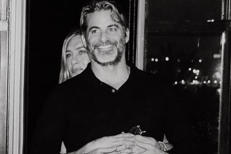 futemax link - Homem de camiseta preta sorri enquanto mulher atrás dele o abraça pela cintura. Ambiente escuro com janela mostrando luzes noturnas ao fundo.