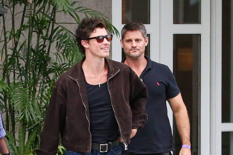 videospon - Dois homens caminham lado a lado em calçada próxima a edifício com portas de vidro. Homem à frente usa jaqueta marrom, camiseta preta e óculos escuros; homem atrás veste camiseta polo preta e calça escura.