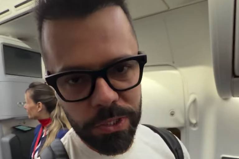 palmeiras x botafogo-sp palpites - Homem de barba e óculos escuros usa camiseta branca e mochila preta dentro da cabine de um avião. Ao fundo, uma mulher está sentada próxima à janela.
