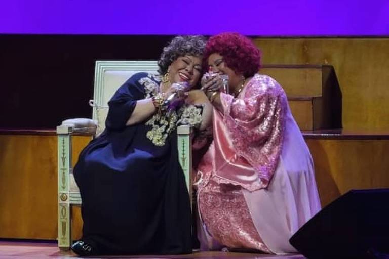 Sexo foda com mina rabuda bem gostosa - Duas mulheres negras sentadas no palco, próximas uma da outra, cantando em um microfone compartilhado. Uma veste roupa preta e a outra um vestido rosa. Fundo roxo com texto 'VOZES NEGRAS' e 'Momento icônico!' acima delas.